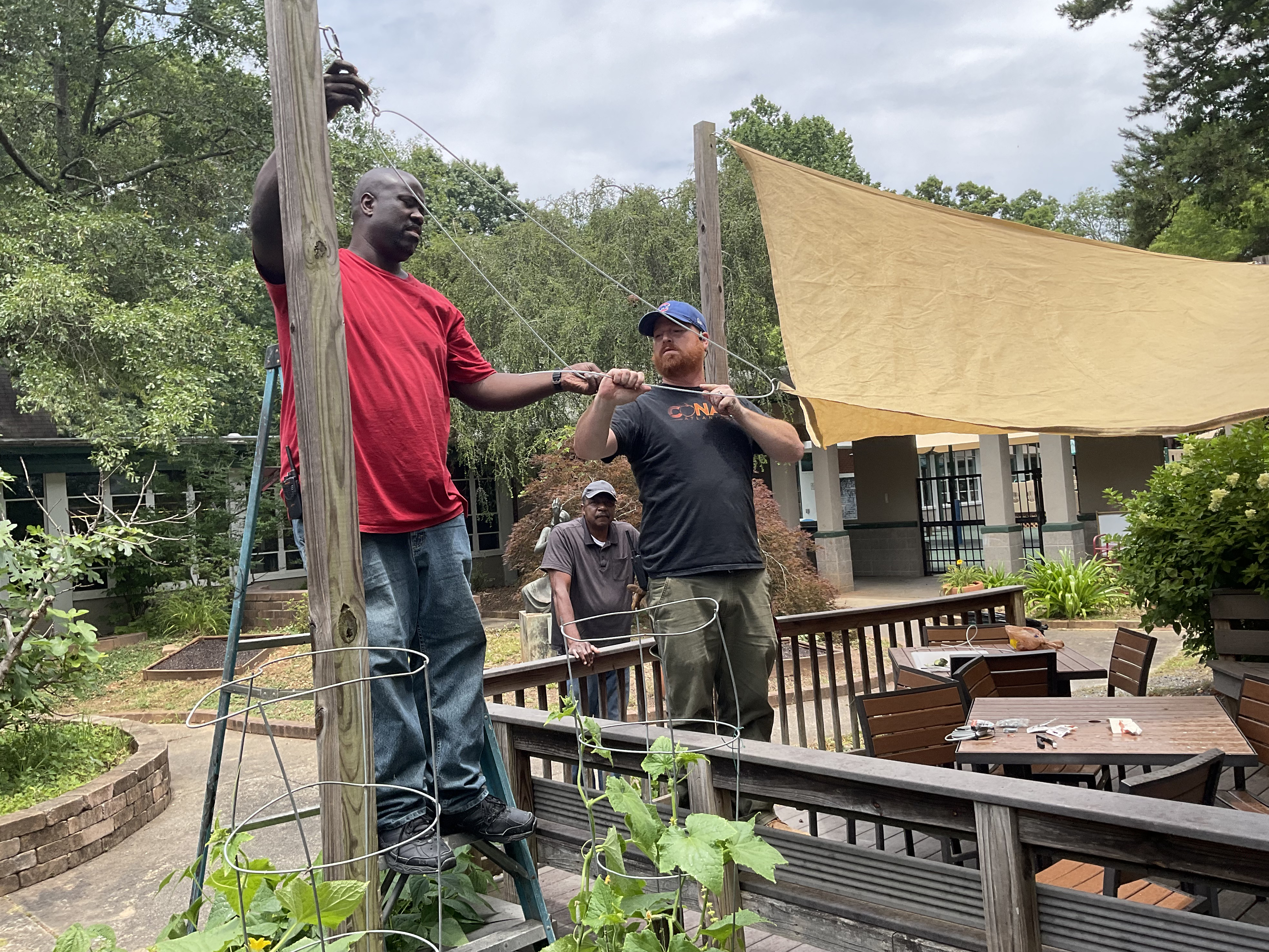Image: Two men repair a canopy over an outdoor patio while another looks on.