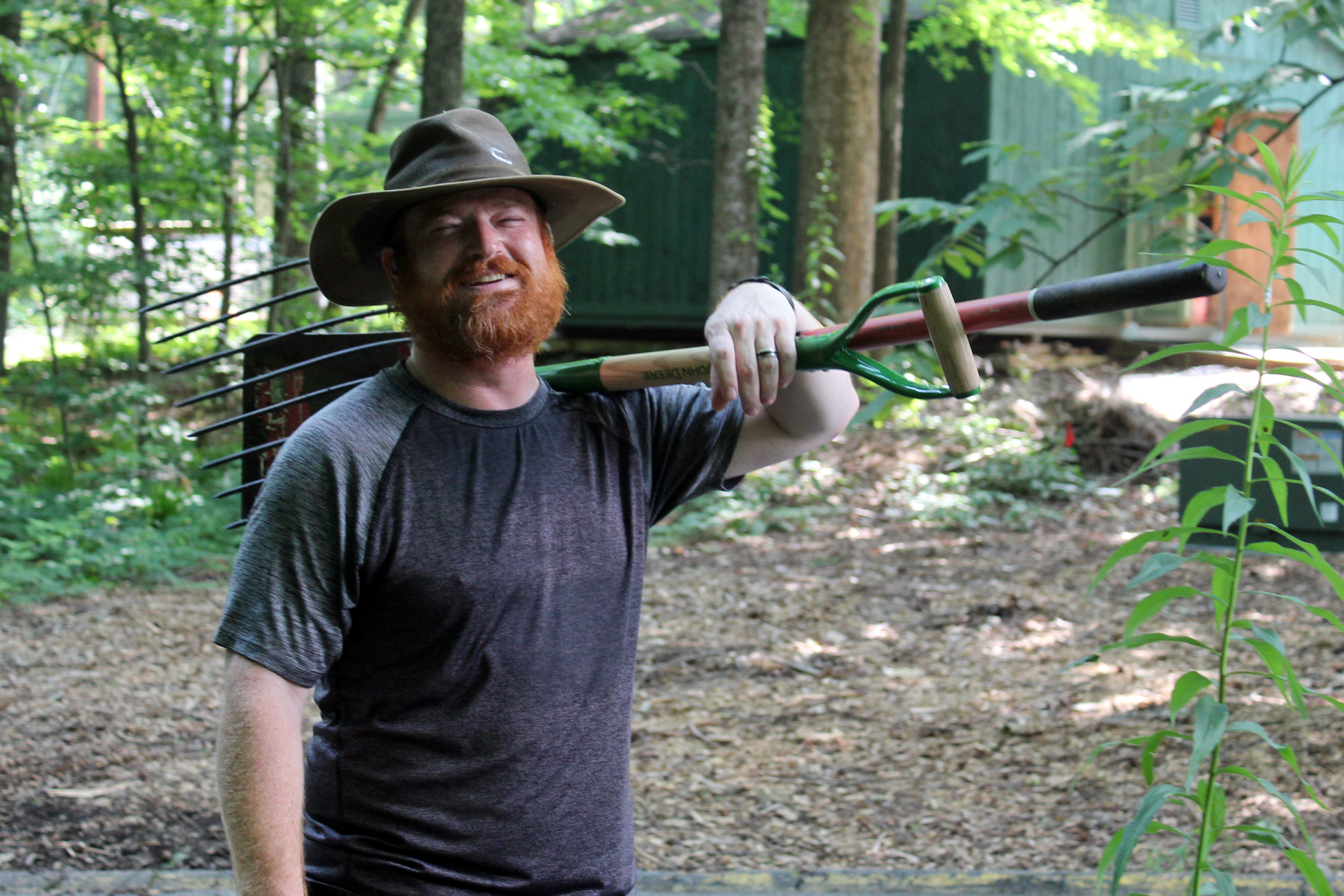 Image: A smiling man wearing a brimmed hat holds a shovel over his shoulder