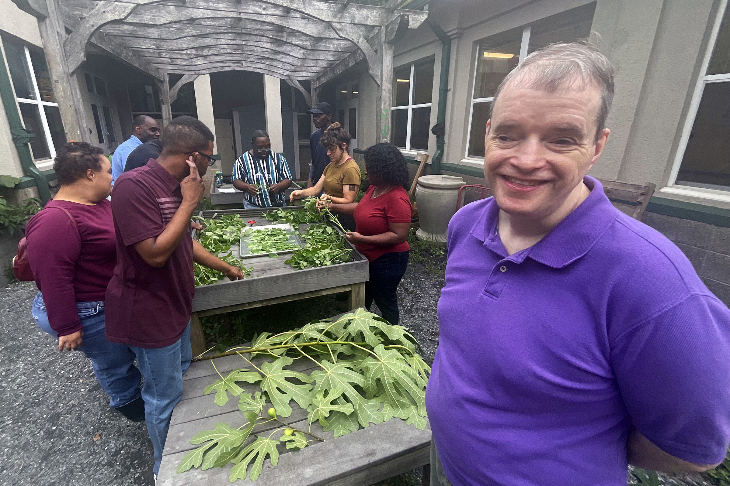 Justin (R) smiles for the camera while Horticultural Therapist Lauren Ladov leads an activity in the courtyard garden with several adult participants and June, one of our Direct Support Professionals.