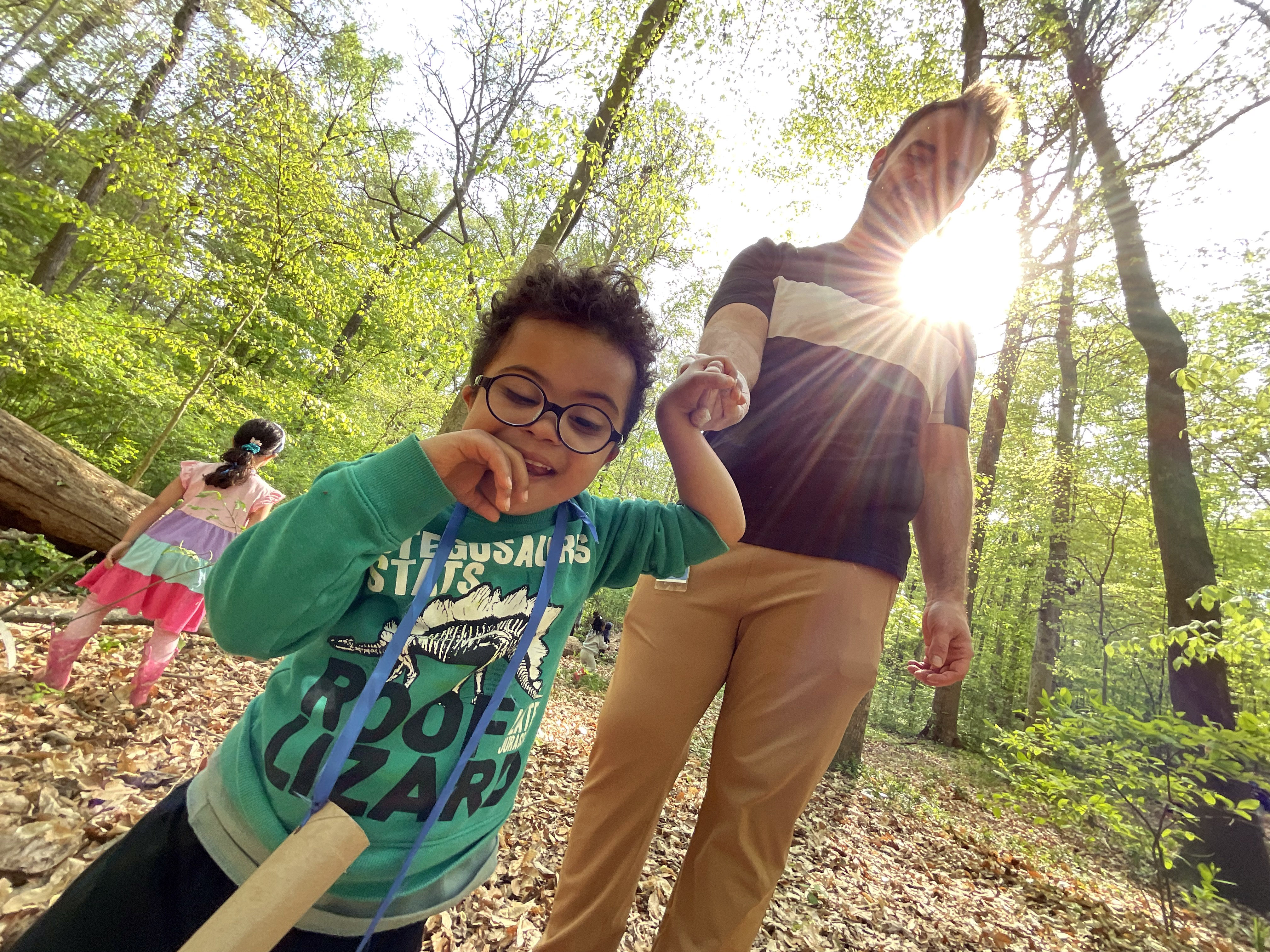 Image of a child and adult in Frazer Forest, holding hands, smiling at the camera, with the sun beaming through trees behind them